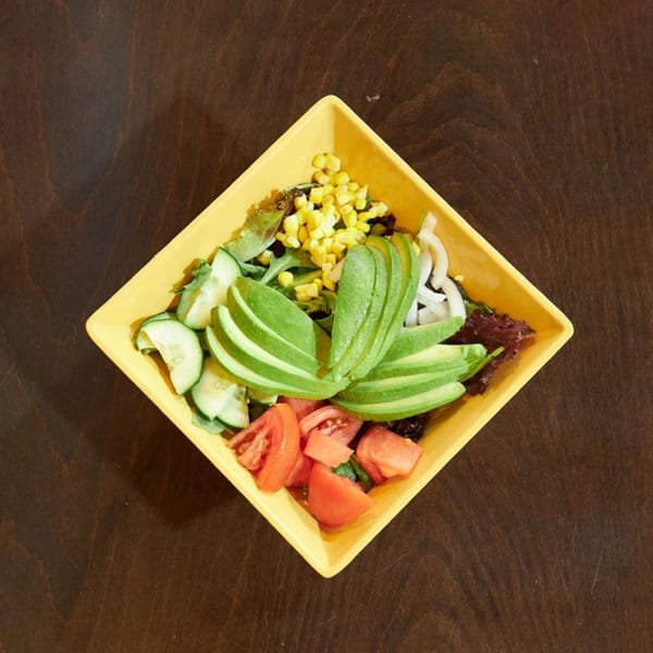 A vibrant salad in a yellow square bowl, featuring avocado, corn, tomatoes, cucumbers, and onions.