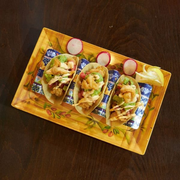 Three Mis Taquitos shrimp tacos with guacamole, sauce, and radishes on a decorative yellow plate.