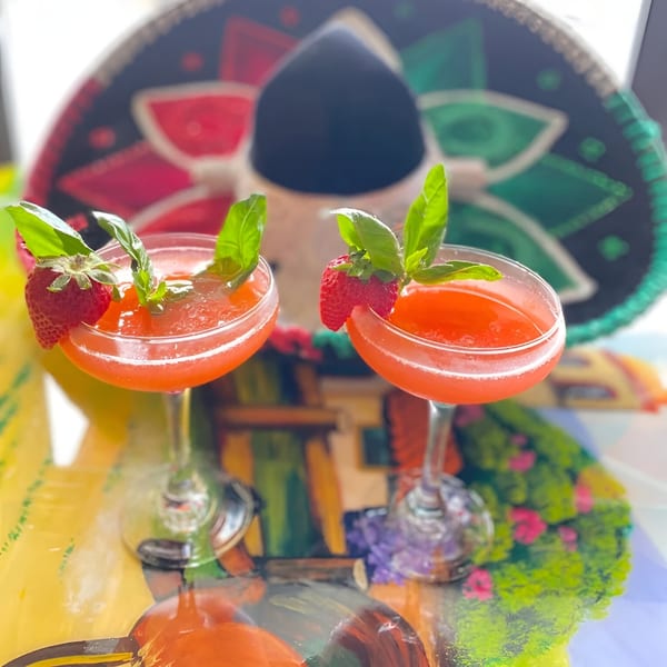Two strawberry margaritas garnished with basil and strawberries, with a sombrero in the background.